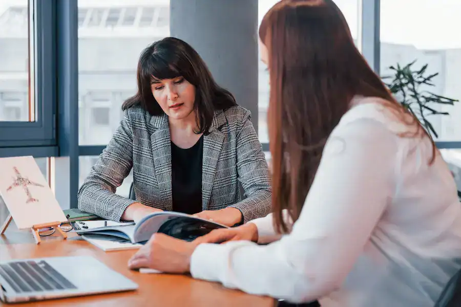 Two women are sitting at a table in an office setting, looking at a document together. One woman is wearing a checked blazer, and the other is in a white blouse.