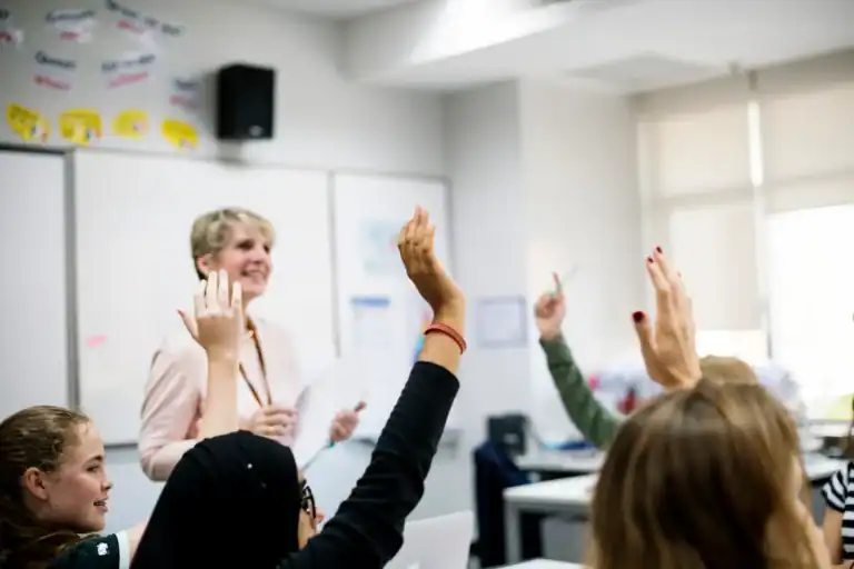 A classroom scene with multiple students raising their hands while a smiling teacher stands at the front holding papers.